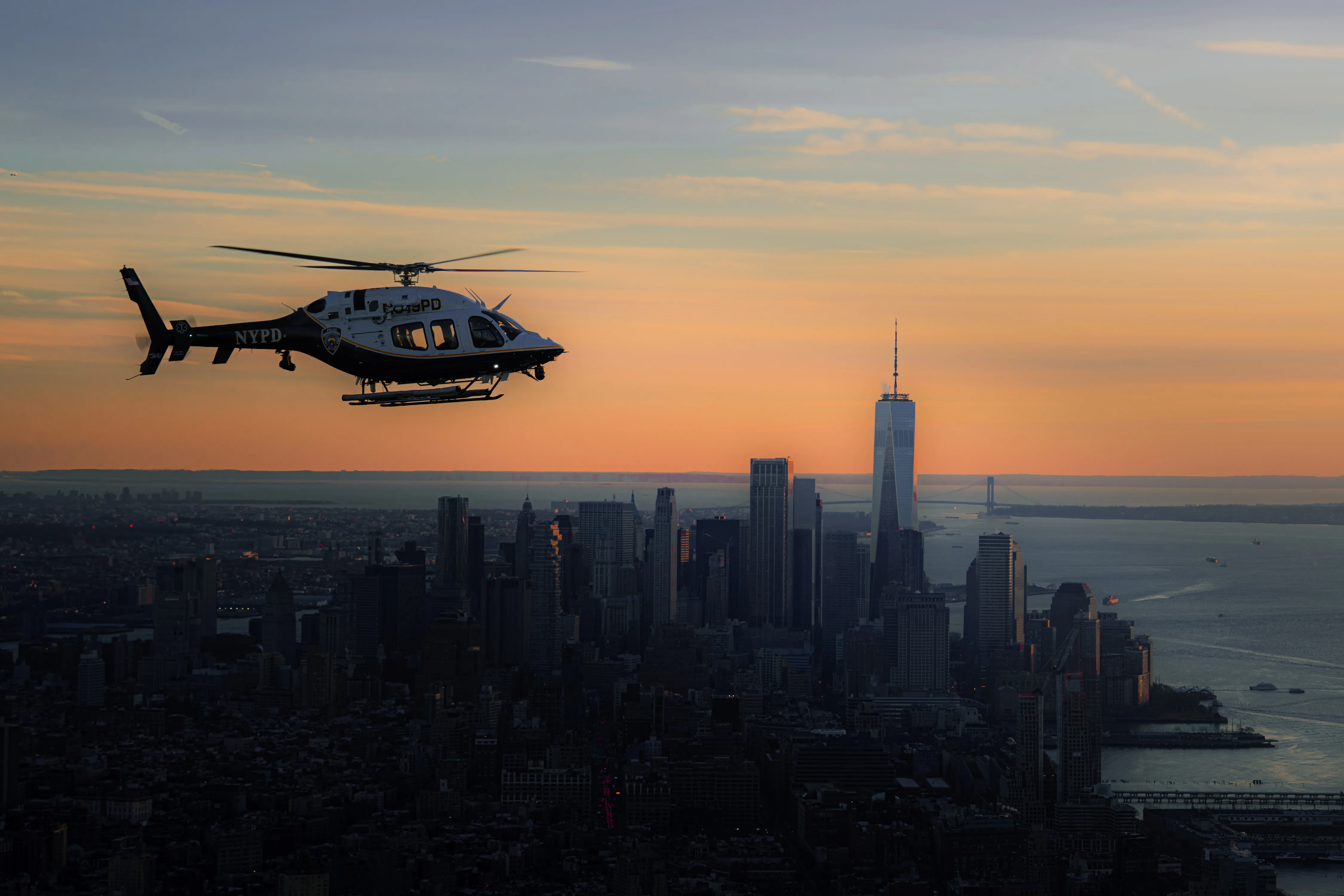A helicopter hovering over Manhattan with One World Trade Center against an orange sky